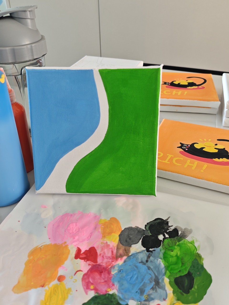 Blue and green acrylic paint on an entirely hand-painted unfinished canvas beside a colorful worktable palette